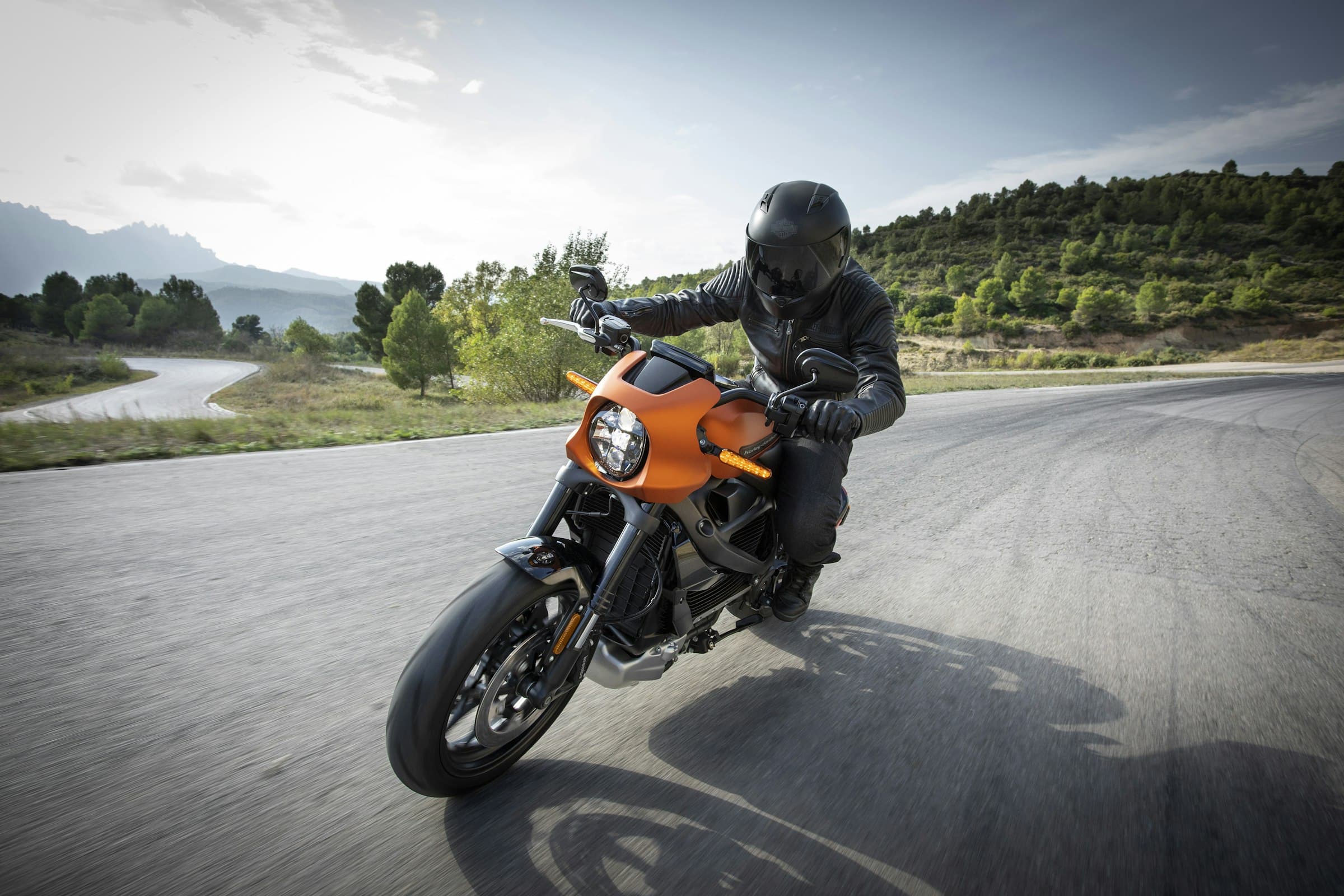 Rider in black leathers leaning an ember-orange motorcycle through a mountain corner